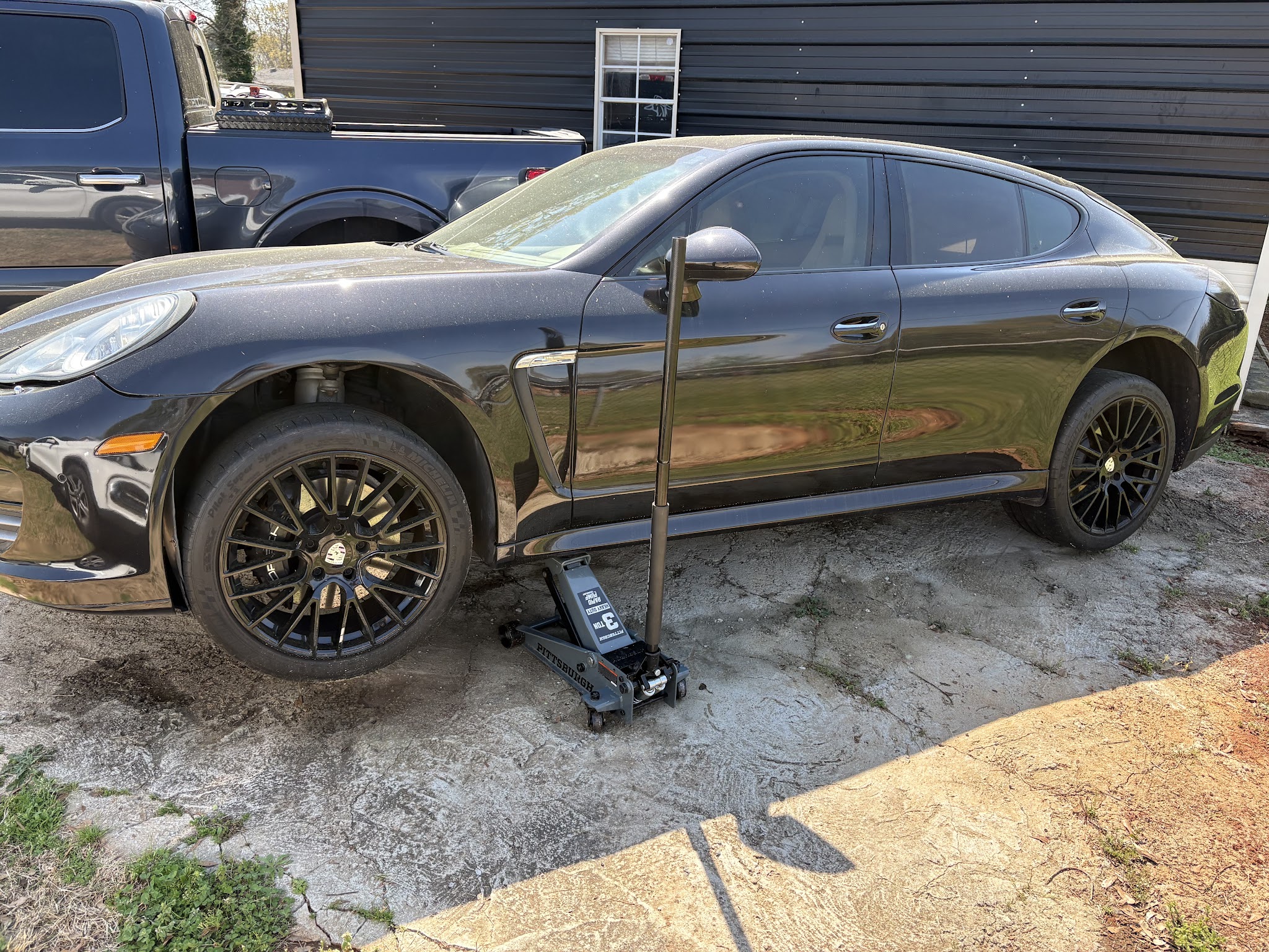 A black Porsche Panamera with a front wheel off, on a floor jack in a driveway