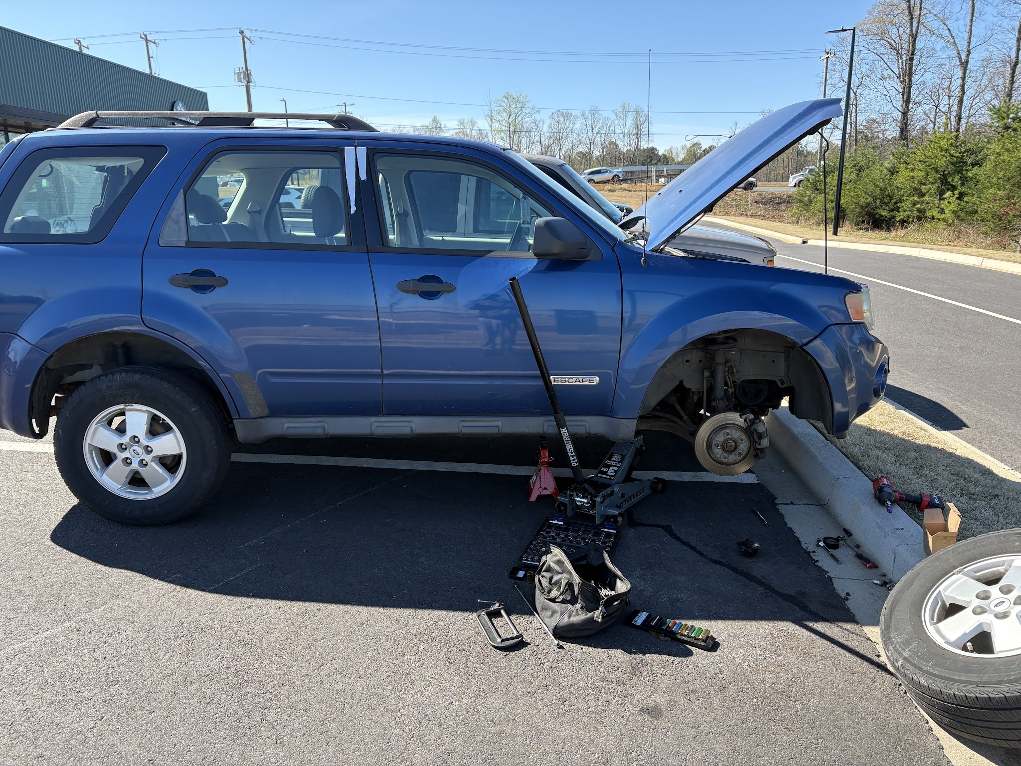 A blue SUV with the hood up and a wheel off, being worked on in a car park